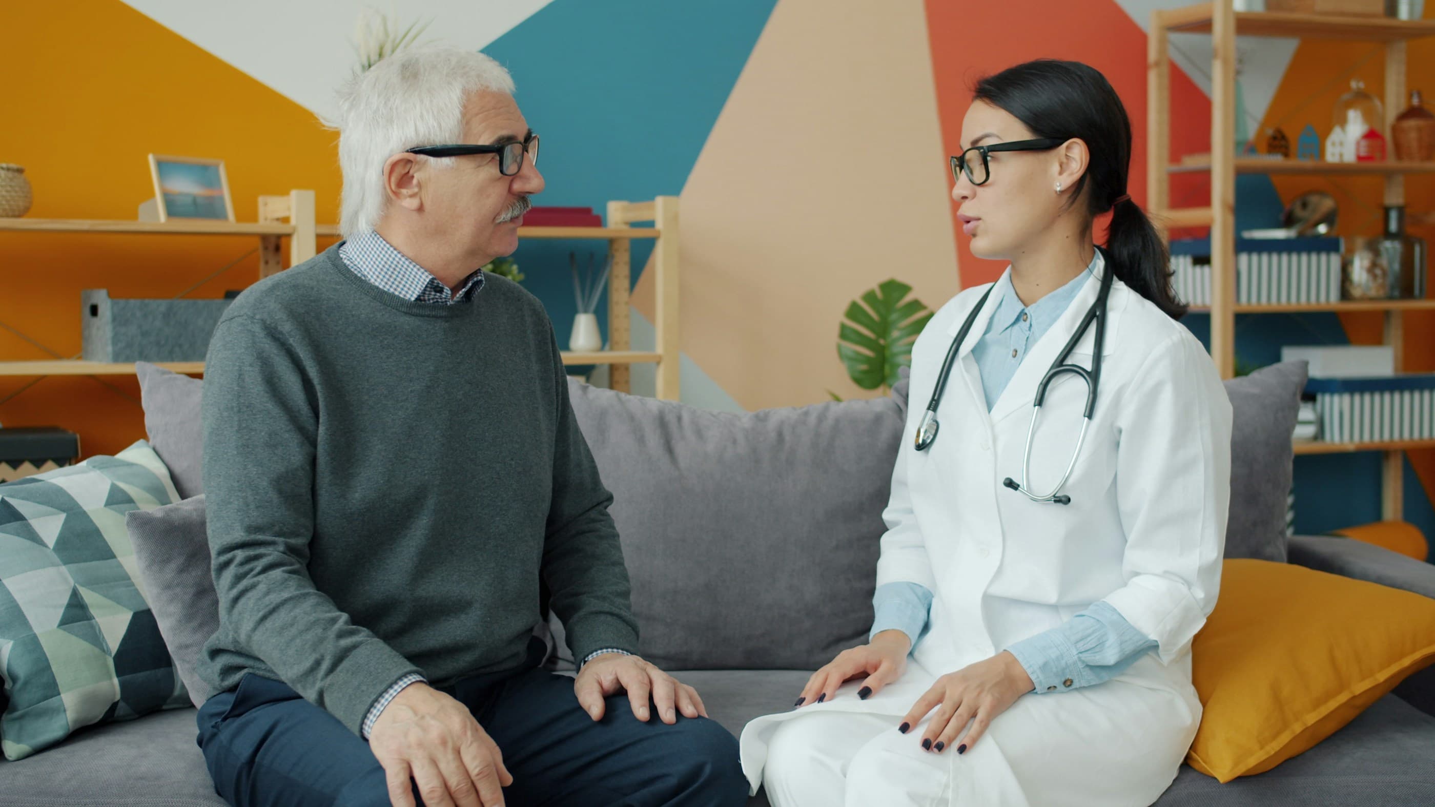 A physician and an older patient sit on a sofa, talking in a bright, informal room