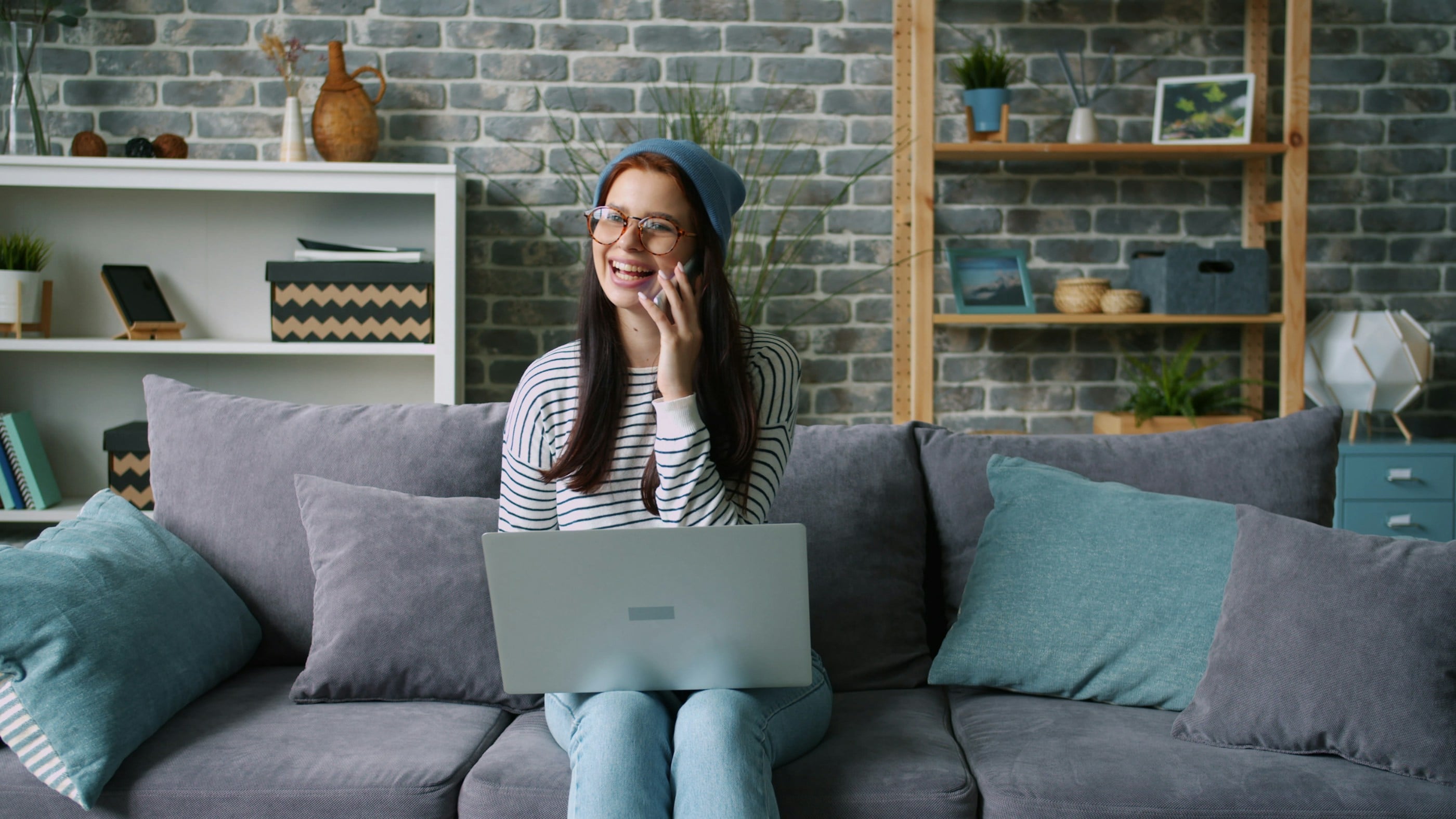 A woman sits on a sofa at home with a laptop on her knees, talking on the phone